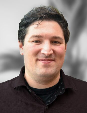 Dr. Jeremy Ford, an Optical Scientist responsible for the design and integration of complex optical systems, smiles at the camera with his short dark hair and dark button-up shirt against a blurred background.