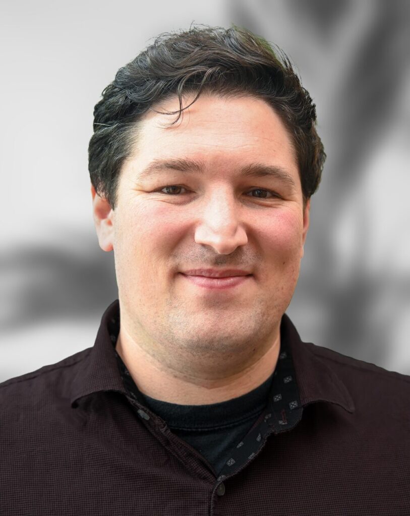 Dr. Jeremy Ford, an Optical Scientist responsible for the design and integration of complex optical systems, smiles at the camera with his short dark hair and dark button-up shirt against a blurred background.