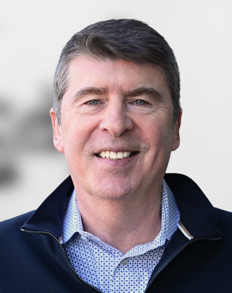 A middle-aged man with short brown hair, Lee McGrath brings more than twenty years of financial leadership experience to his role as Chief Financial Officer, smiling at the camera in a dark jacket over a patterned shirt against a plain light background.