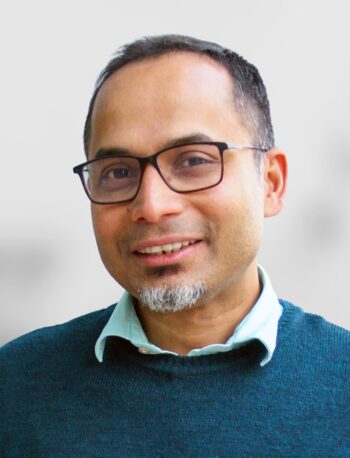 A photo of Partha Deb, a man with short hair, glasses, and a goatee is smiling, wearing a teal sweater over a light blue collared shirt, in front of a plain light background.