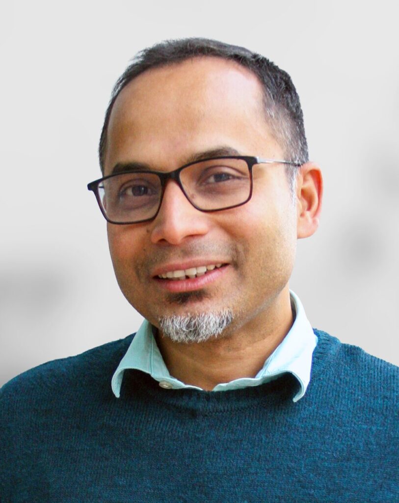 A photo of Partha Deb, a man with short hair, glasses, and a goatee is smiling, wearing a teal sweater over a light blue collared shirt, in front of a plain light background.