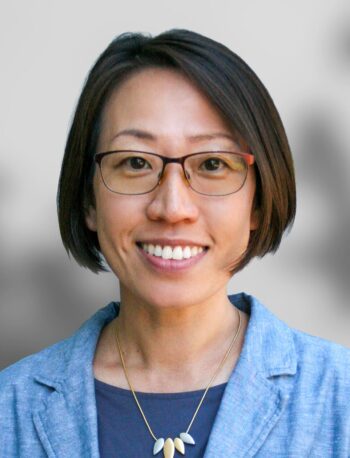 A woman with short brown hair and glasses, wearing a blue blazer, a dark top, and a necklace, smiles at the camera against a blurred background.