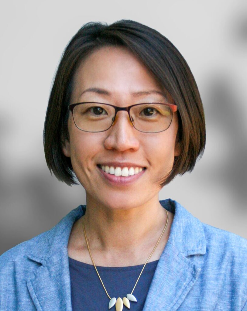 A woman with short brown hair and glasses, wearing a blue blazer, a dark top, and a necklace, smiles at the camera against a blurred background.