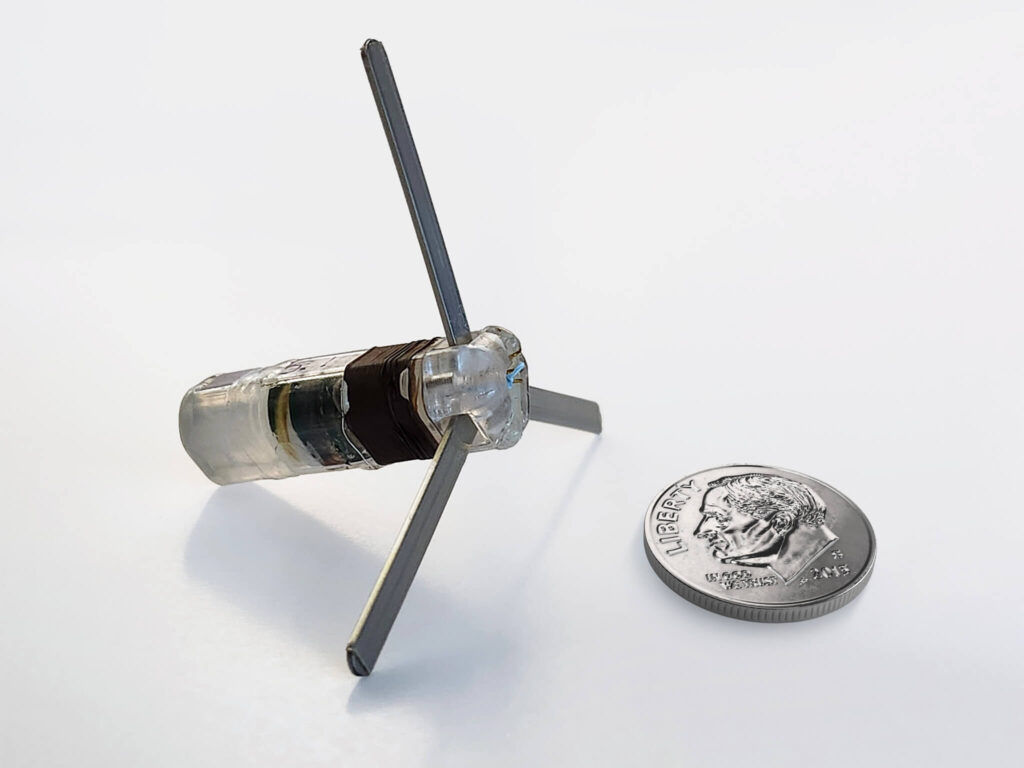 A small robotic device with three extended legs is shown next to a U.S. dime for scale, all on a white background.