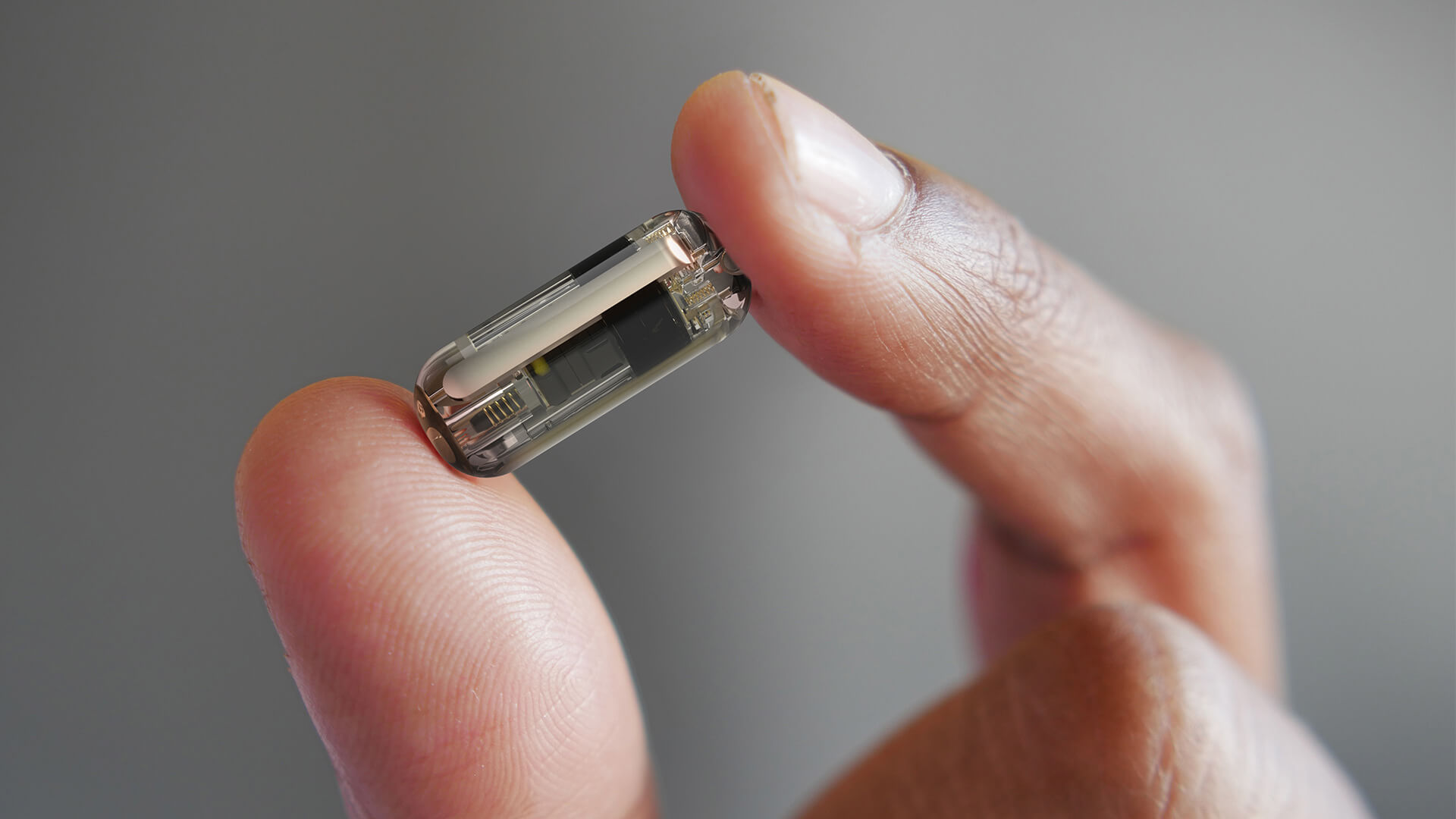A hand holds a small, transparent electronic device between two fingers against a blurred gray background.