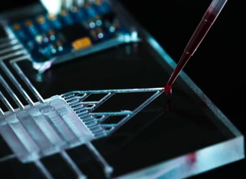 A pipette dispensing a red liquid onto a microfluidic chip with electronic components visible in the background.