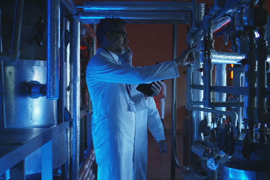 Two scientists in lab coats inspect industrial pipes and equipment under blue lighting in a laboratory or facility.