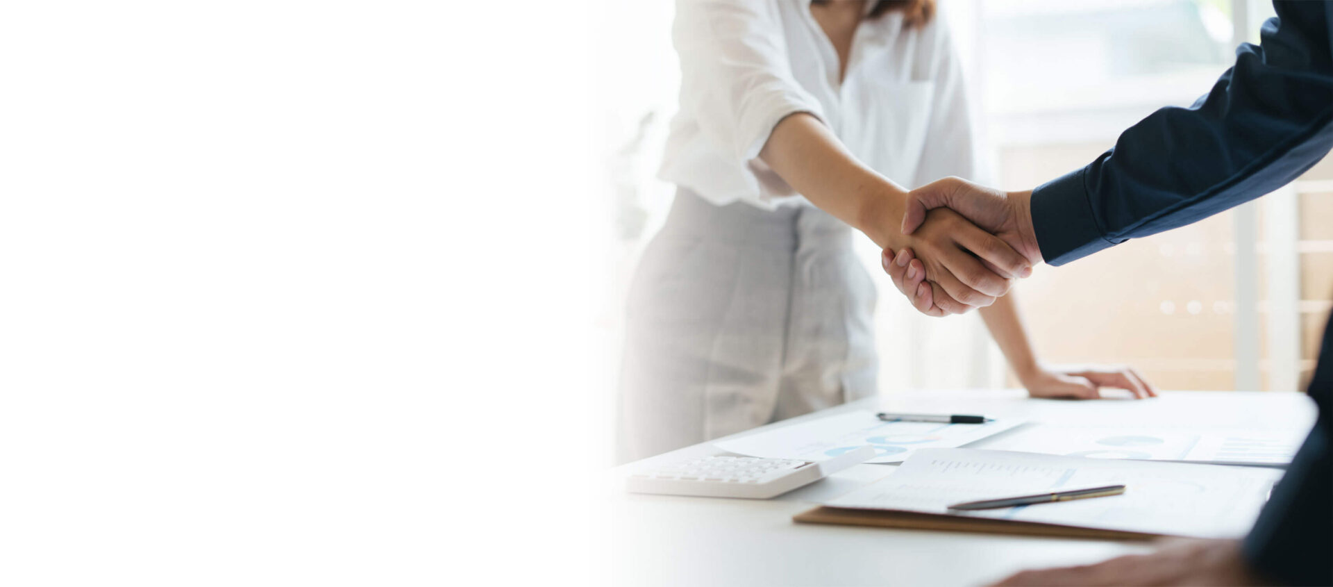 Two people in business attire shake hands across a desk with documents, a calculator, and a pen visible.