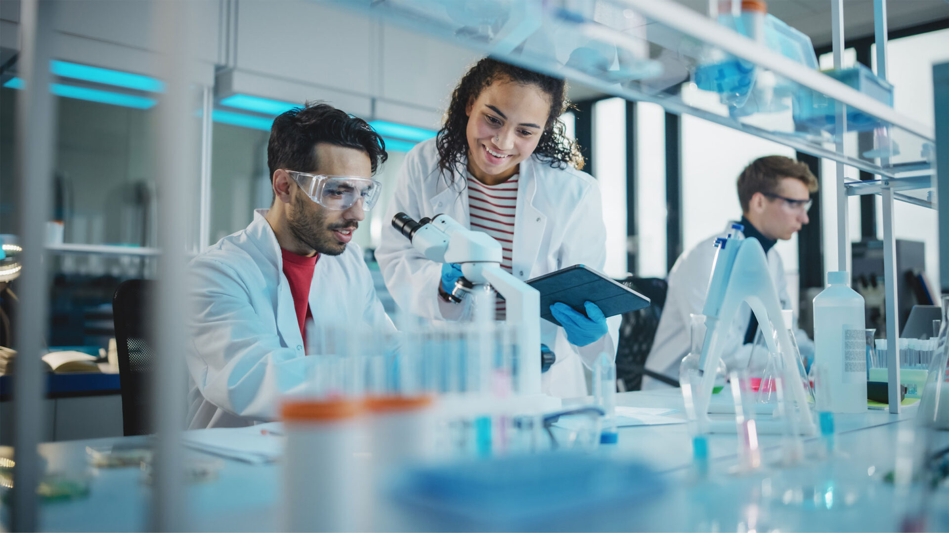 Two scientists in lab coats work together at a laboratory bench with test tubes and a microscope; one holds a tablet while another scientist works in the background.